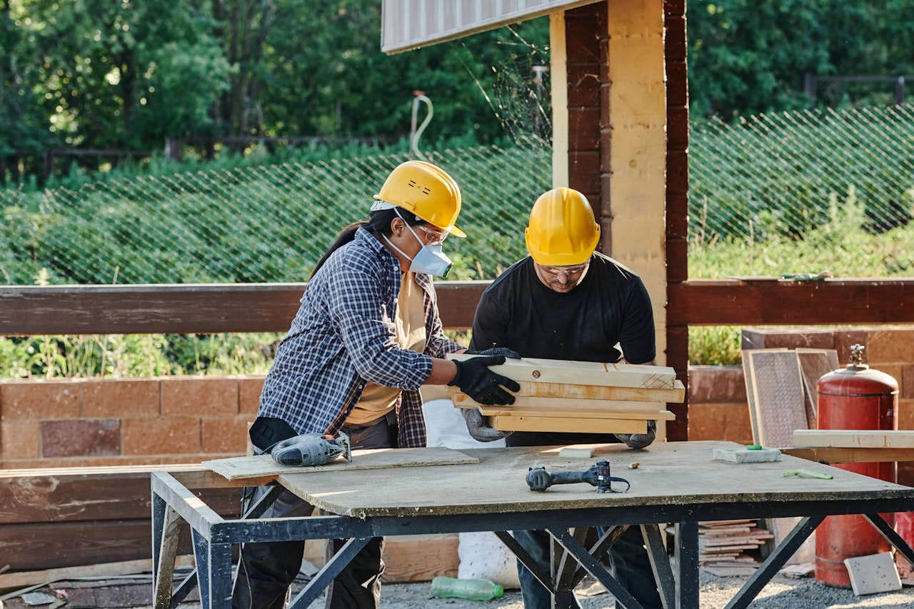 Two construction workers organize wooden planks on a table at a sunny outdoor site.